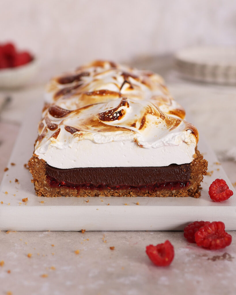 Smore tart cut on a white board showing the layers of crumb base, raspberry jam, chocolate ganache and fluffy meringue.