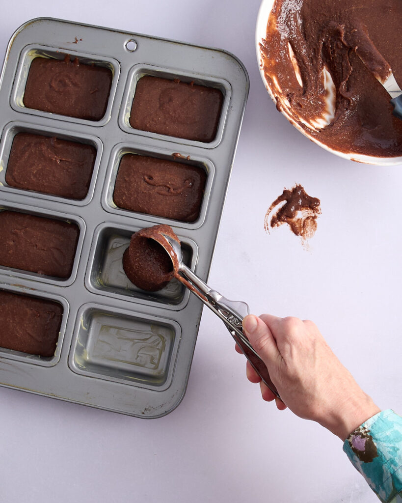 Sandra scooping chocolate financier batter into a mini loaf pan, with a mixing bowl and smears of batter on the bench nearby.