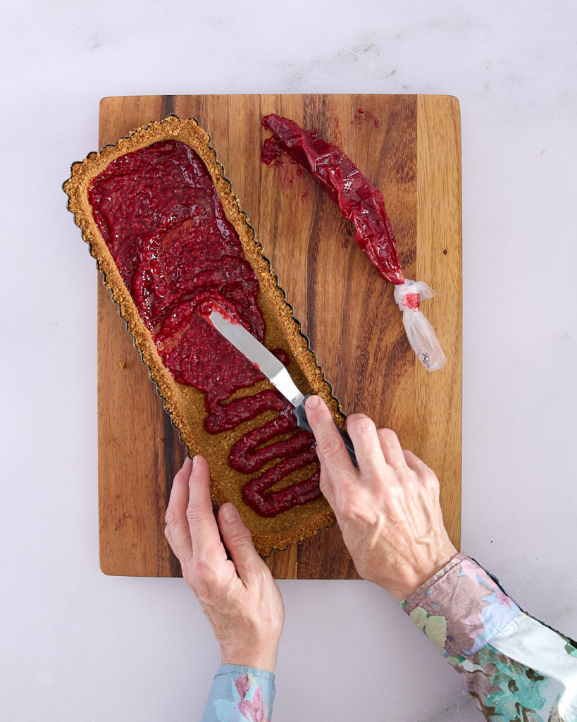 Sandra spreads raspberry jam with an offset spatula inside a rectangular tart crust, with a piping bag of jam to the side.