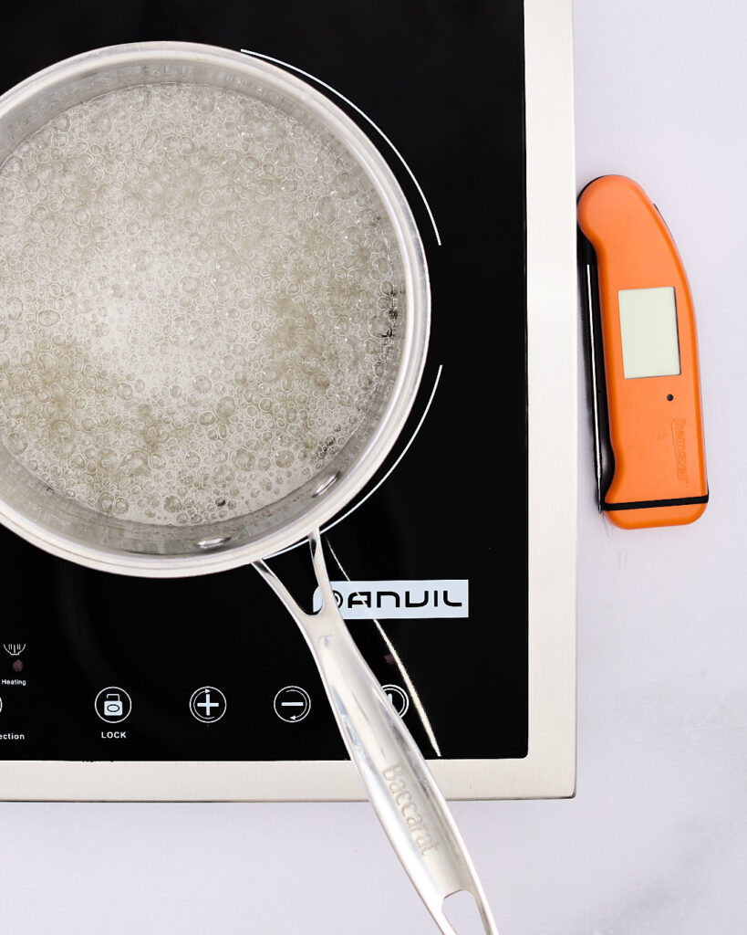 A saucepan of bubbling sugar syrup on an induction cooktop, shown mid-boil with a thermometer alongside—step in making marshmallow.
