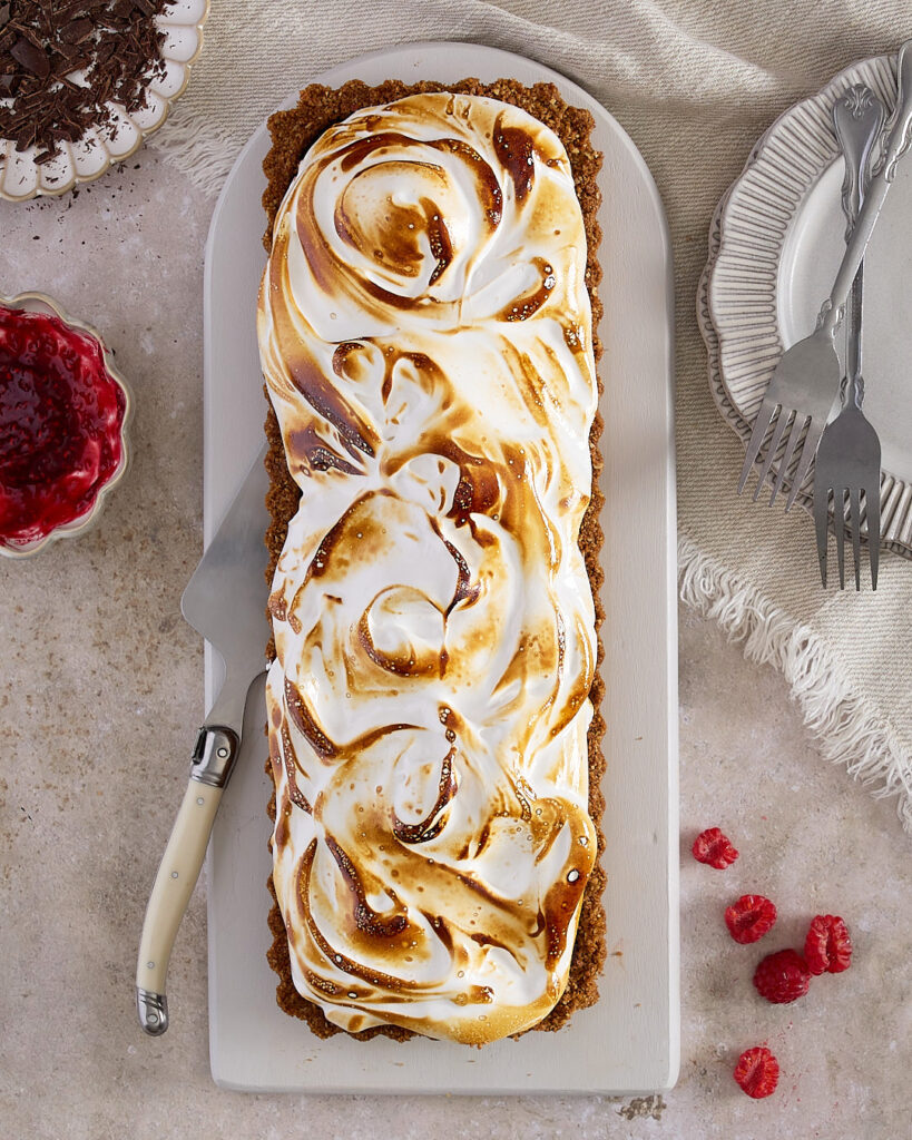 A whole chocolate raspberry marshmallow tart topped with toasted swirls of homemade marshmallow, set on a white platter with plates, forks, and raspberries scattered nearby.