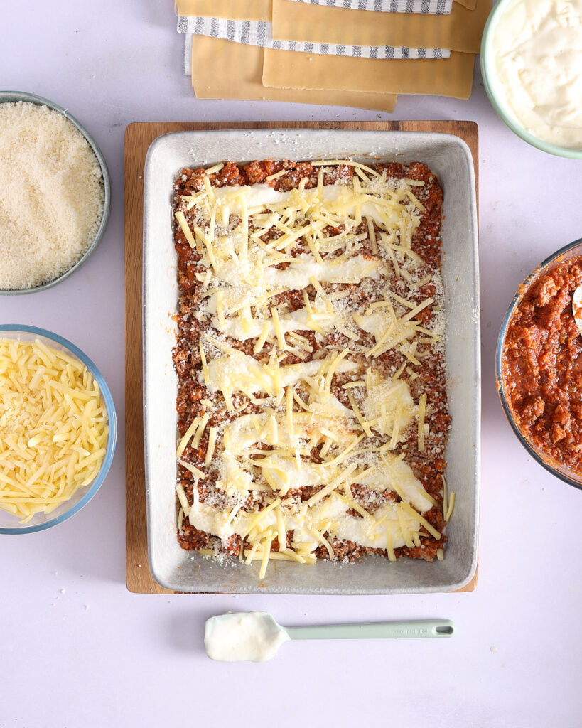 Layering béchamel, shredded mozzarella, and grated Parmigiano Reggiano cheese over the bolognese sauce.