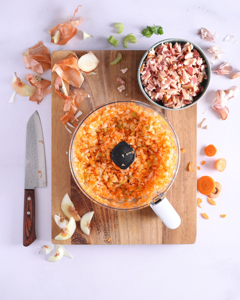 Food processor roughly chopped carrot, celery, and onion with diced bacon to the side.