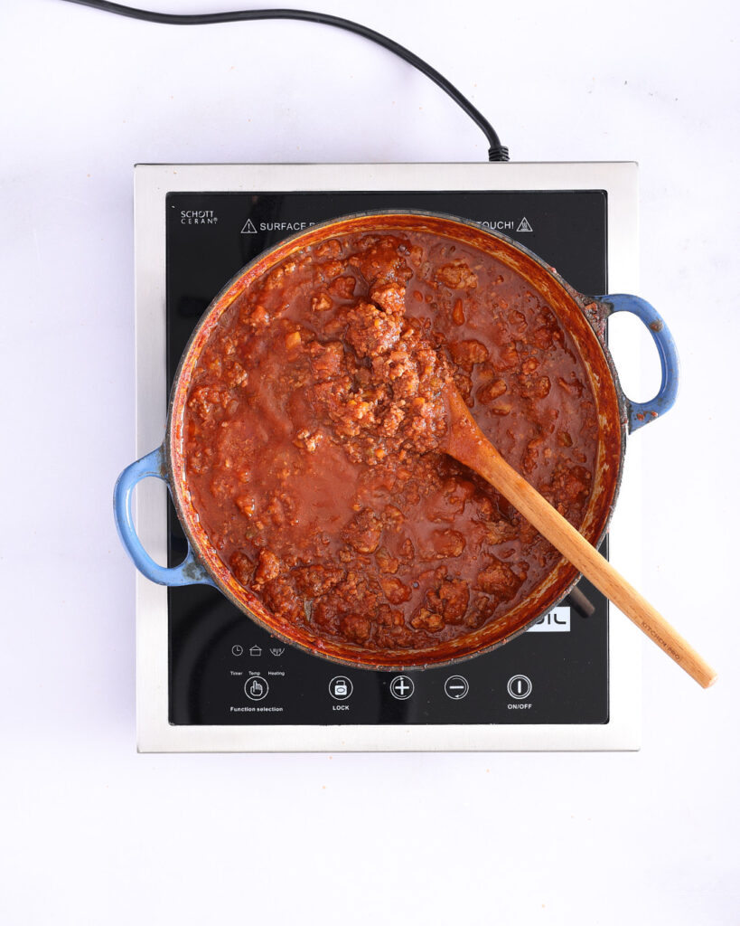 Rich red bolognese sauce simmering, dotted with herbs and visible chunks of meat and vegetables.