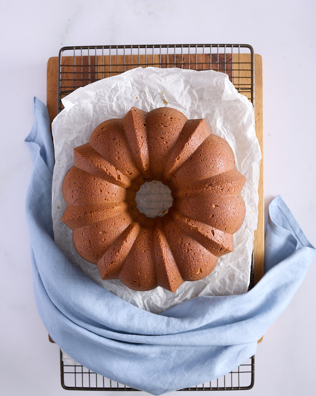 Lemon blueberry bundt cake released from its pan, cooling upside down on a wire rack lined with baking paper, draped with a blue napkin.