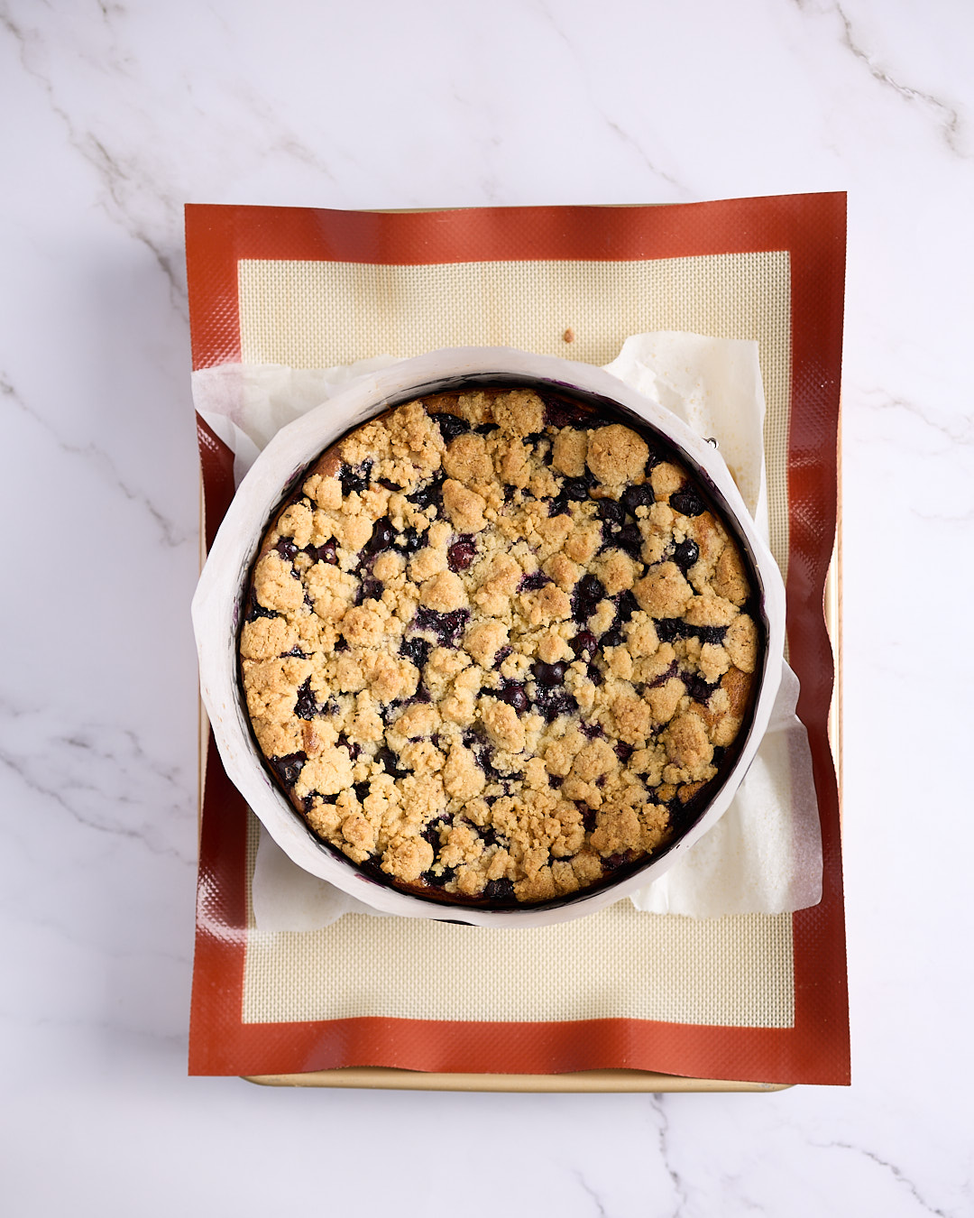 Overhead image of a freshly baked blueberry rosemary crumble cheesecake in a springform pan on a silicone-lined tray, showing a golden crumb and set edges.