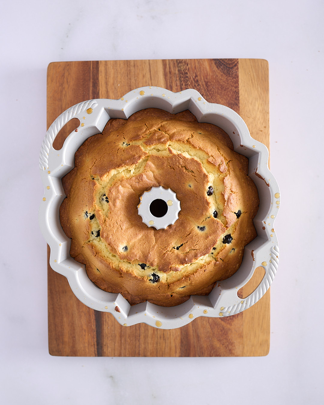 Baked lemon blueberry bundt cake still in its pan, golden brown with blueberries visible, resting on a wooden board after coming out of the oven.