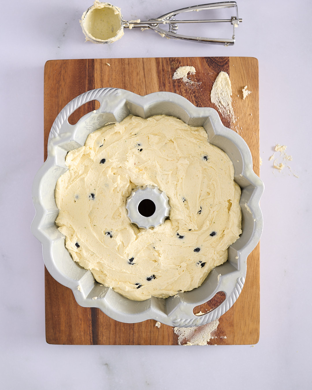 Bundt pan filled with pound cake batter dotted with fresh blueberries, smoothed with an offset spatula; ice cream scoop resting nearby on a wooden board—step showing how to layer and fill batter for a lemon blueberry rosemary bundt cake.