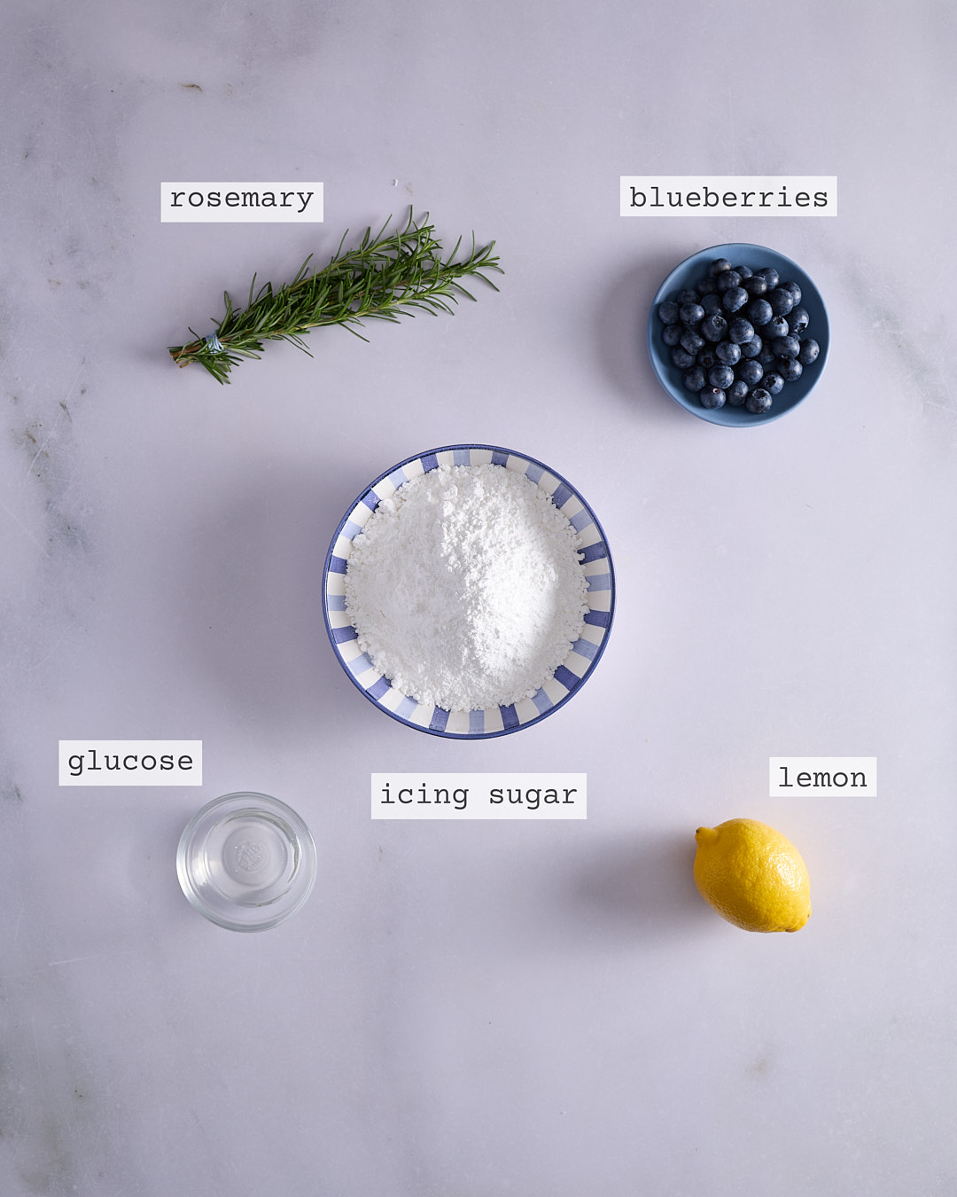 Overhead of icing ingredients for lemon blueberry rosemary bundt cake including rosemary, blueberries, icing sugar, lemon, glucose, and granulated sugar.