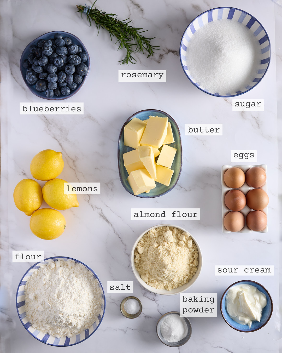 Flat lay of ingredients for lemon blueberry rosemary bundt cake showing lemons, blueberries, rosemary, butter, sugar, eggs, almond flour, sour cream, flour, salt, and baking powder.