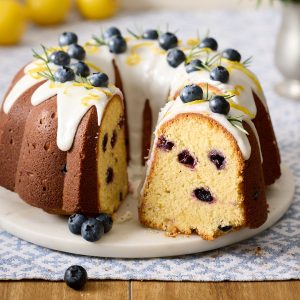 Lemon blueberry rosemary bundt cake with white lemon glaze, decorated with fresh blueberries, lemon zest, and rosemary sprigs, sliced to show moist crumb.