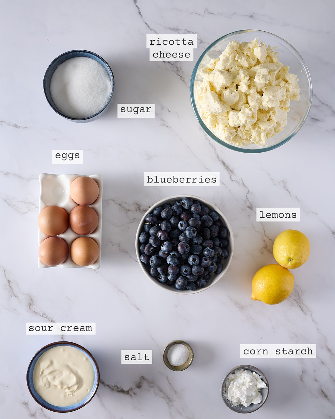 Overhead photo of labelled cheesecake ingredients for a low sugar blueberry ricotta cheesecake recipe showing traditional ricotta, sour cream, sugar, blueberries, lemons, corn starch, eggs, and salt arranged on a marble surface.