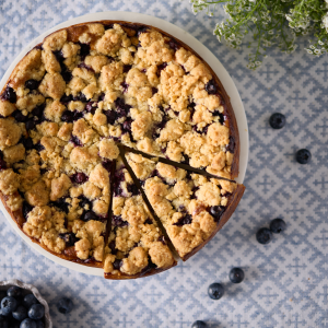 Overhead shot of a blueberry rosemary crumble cheesecake baked in a round tin, topped with golden almond crumble and scattered blueberries, showing the rustic texture and naturally gluten-free crumble topping.