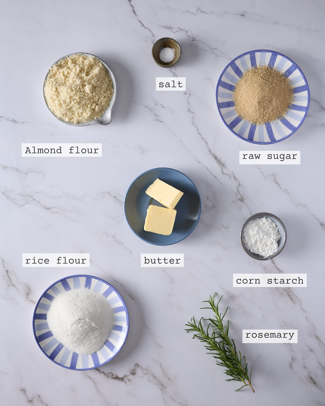Overhead photo of labelled gluten-free rosemary crumble ingredients for a blueberry rosemary crumble cheesecake, showing almond flour, rice flour, corn starch, butter, raw sugar, salt, and a sprig of fresh rosemary on a marble surface.
