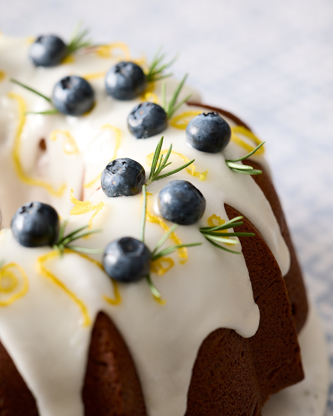 Close-up of a Bundt cake’s surface—glazed top dripping slightly, dotted with blueberries and sprigs of rosemary, lemon zest curling over the edge.