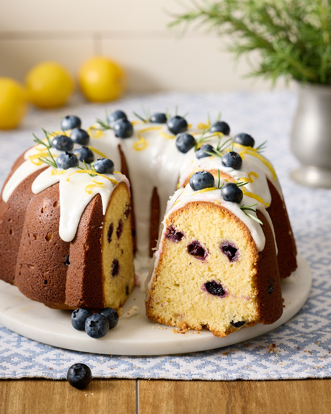 Lemon blueberry rosemary bundt cake with white lemon glaze, decorated with fresh blueberries, lemon zest, and rosemary sprigs, sliced to show moist crumb.