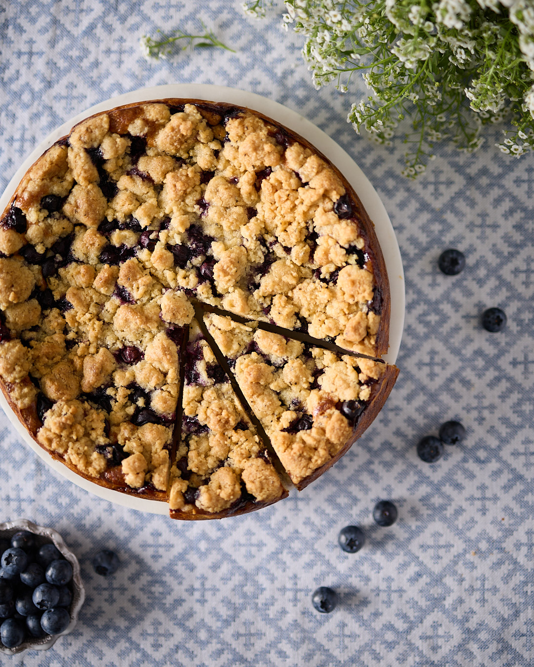 Overhead shot of a blueberry rosemary crumble cheesecake baked in a round tin, topped with golden almond crumble and scattered blueberries, showing the rustic texture and naturally gluten-free crumble topping.
