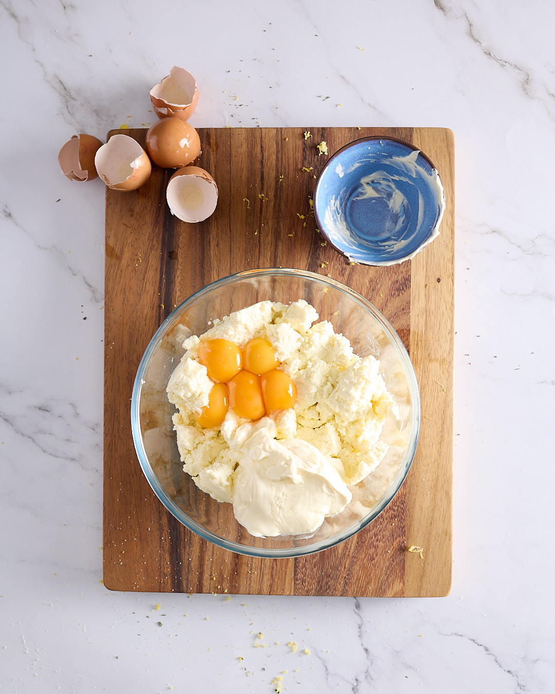 Overhead image showing ricotta, sour cream, and egg yolks in a glass bowl on a wooden board, the start of the low sugar blueberry ricotta cheesecake recipe.