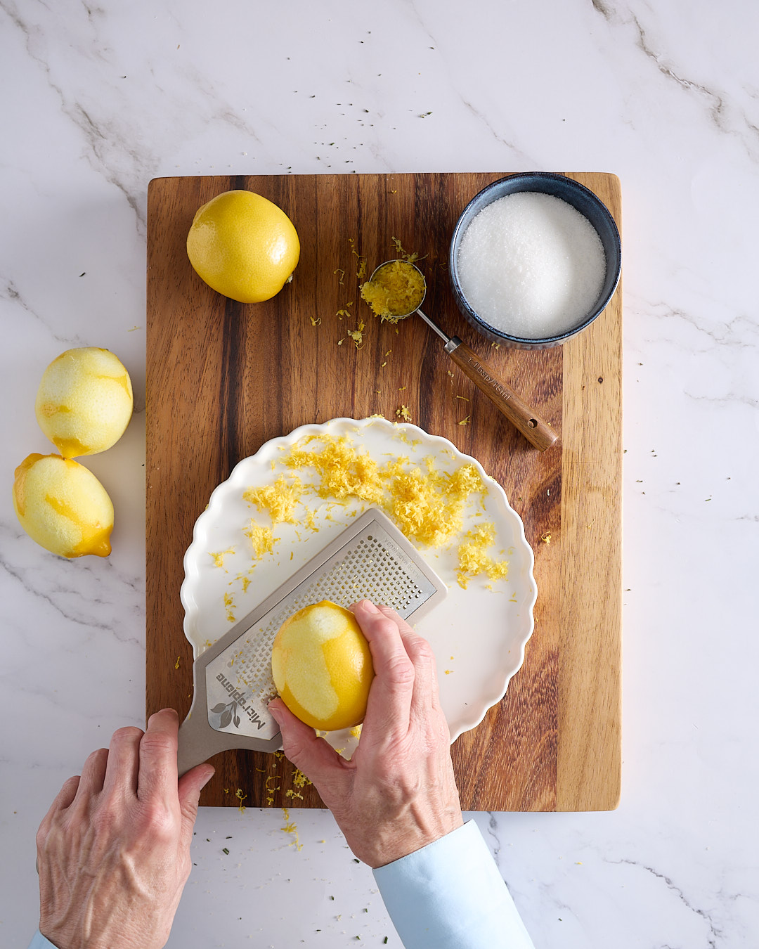 Sandra zesting lemons with a Microplane on a white plate, surrounded by sugar and zest for a blueberry rosemary crumble cheesecake.