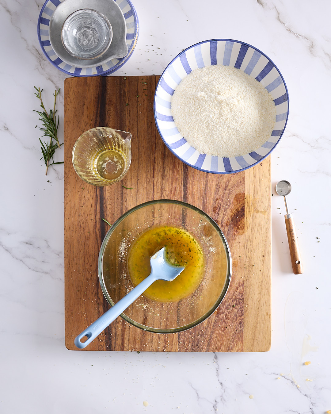 Overhead image showing melted butter infused with chopped rosemary in a glass bowl on a wooden board, part of the preparation for a blueberry rosemary crumble cheesecake.