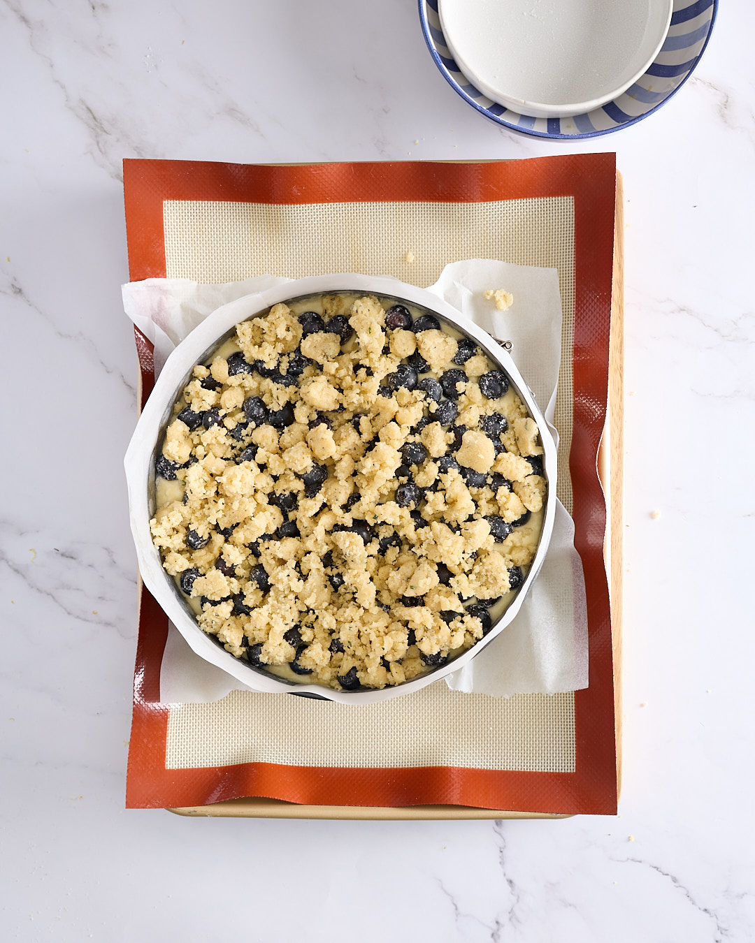 Overhead image of an unbaked easy gluten-free blueberry cheesecake topped with golden rosemary crumble, ready to go into the oven on a silicone-lined tray.