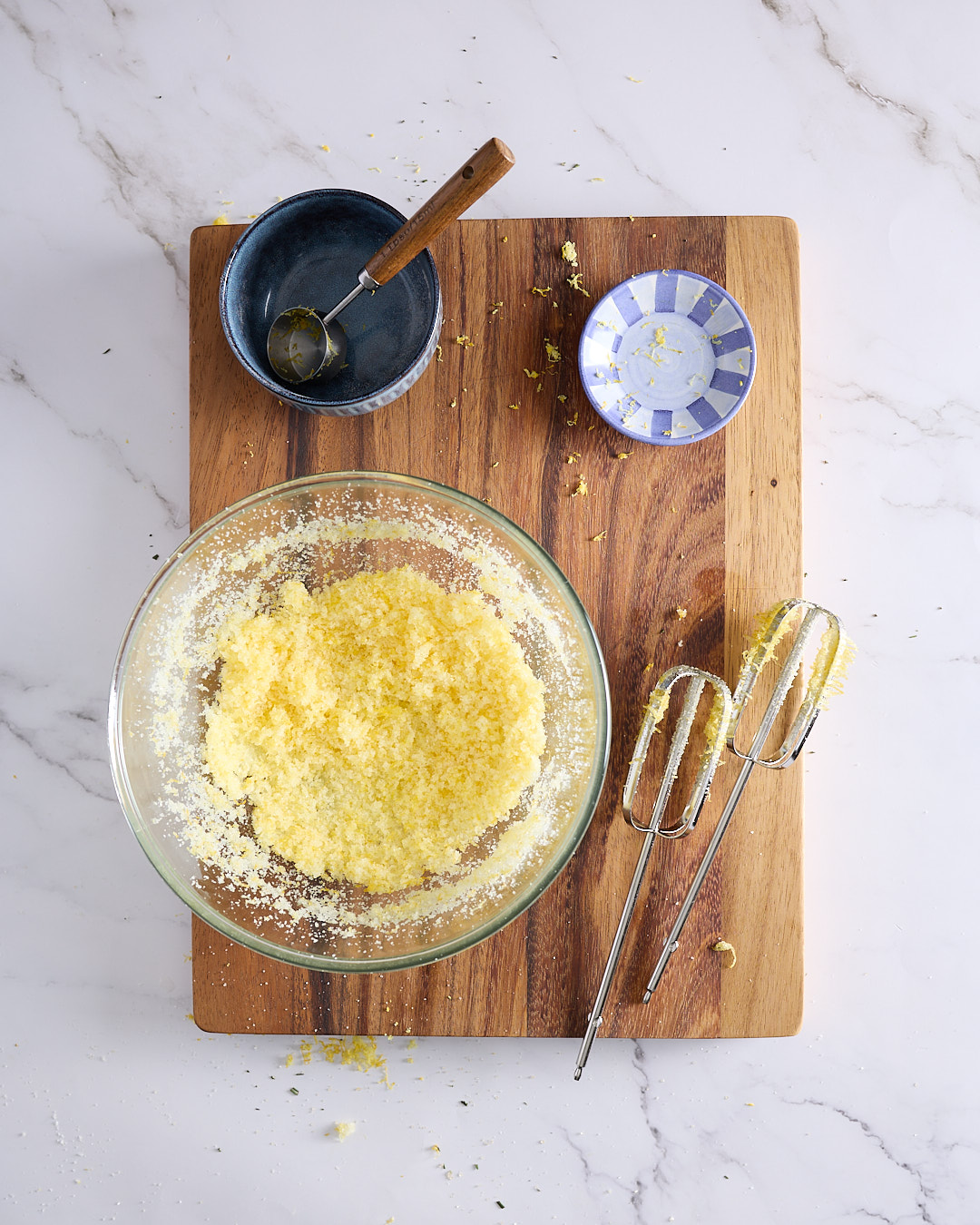 Overhead image of sugar and lemon zest beaten together in a bowl with mixer beaters beside it, showing the start of the low sugar blueberry ricotta cheesecake recipe batter.