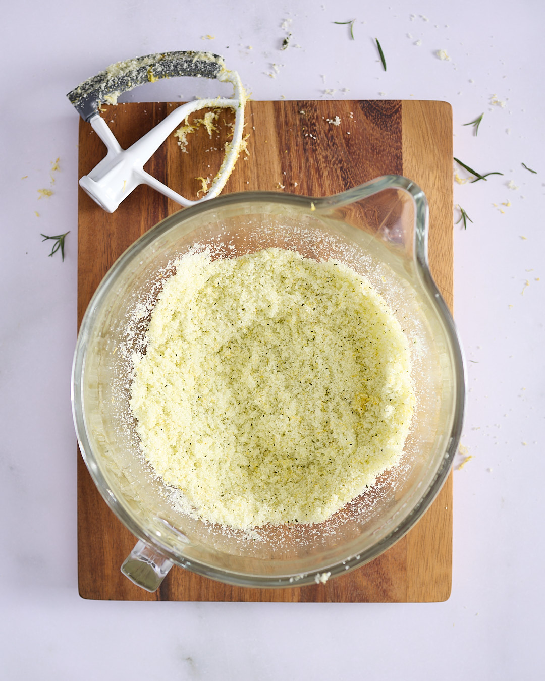 Glass stand mixer bowl filled with granulated sugar mixed with lemon zest and rosemary, paddle attachment resting nearby —before creaming in a lemon blueberry rosemary bundt cake.