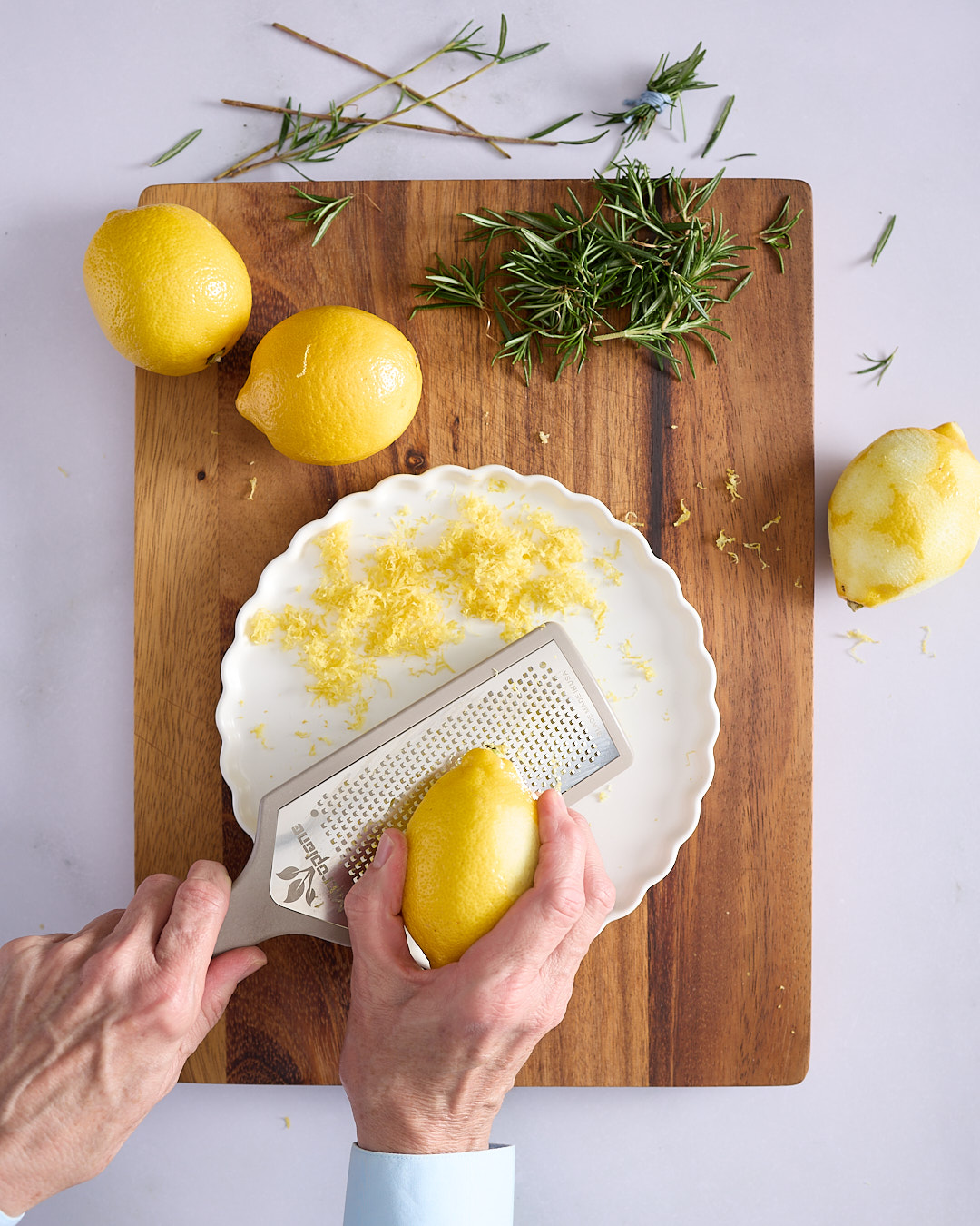 Sandra zesting a lemon with a Microplane over a white plate, with fresh rosemary sprigs and whole lemons on a wooden board—step showing preparation of zest and herbs for a lemon blueberry rosemary bundt cake.