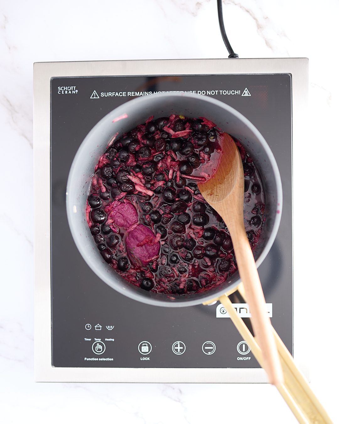 Overhead view of blueberries simmering in a stainless-steel pot on an induction cooktop, with a wooden spoon resting on the side and a bundle of rosemary wrapped in cheesecloth visible among the berries.