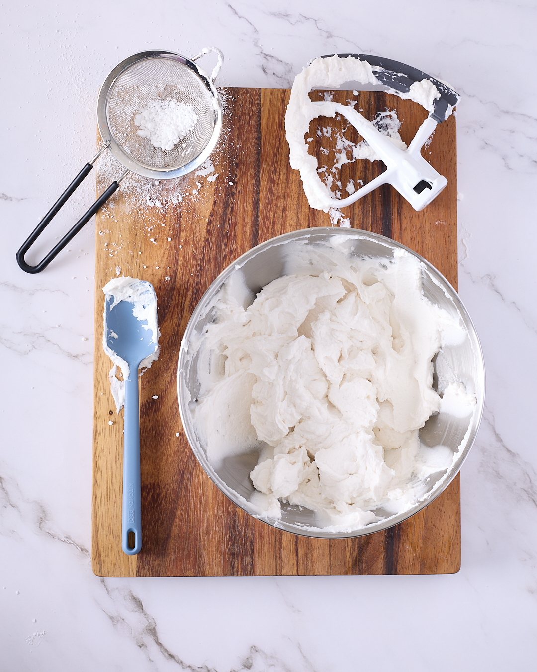 Whipped strained coconut yoghurt in a stainless steel mixing bowl on a wooden board, with a spatula, icing sugar sieve, and stand mixer paddle attachment showing the process of lightly sweetening and aerating the yoghurt.