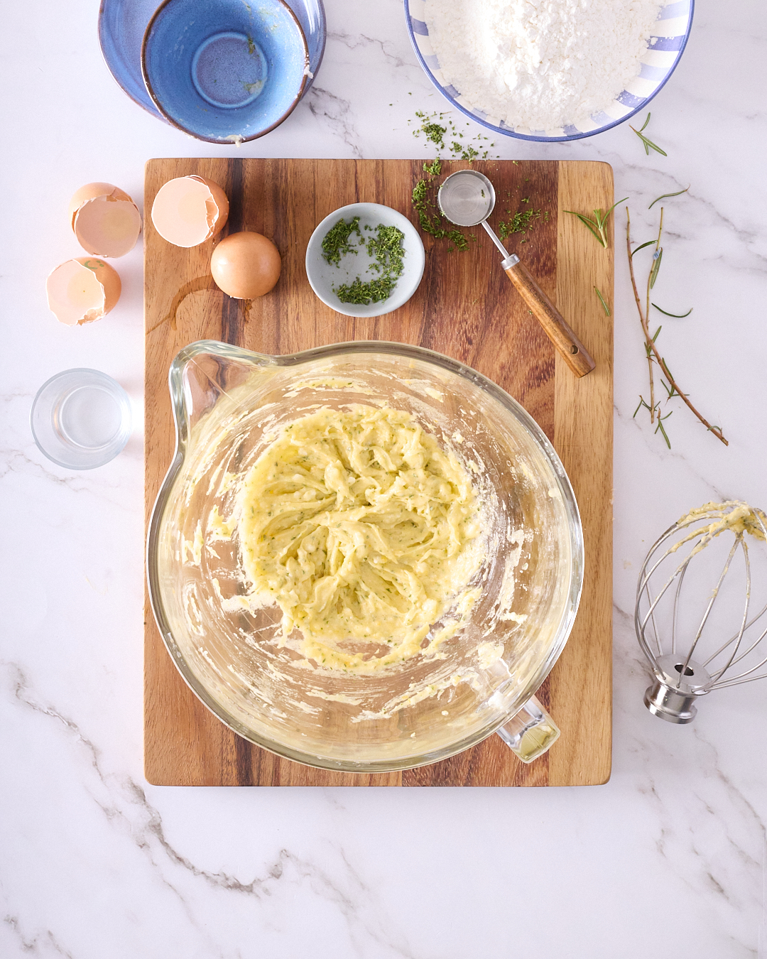 Glass mixing bowl on a wooden board showing creamed egg yolks, coconut oil, and chopped rosemary for gluten free vegan rosemary shortbread, surrounded by bowls of flour, sugar, and rosemary sprigs on a marble countertop.