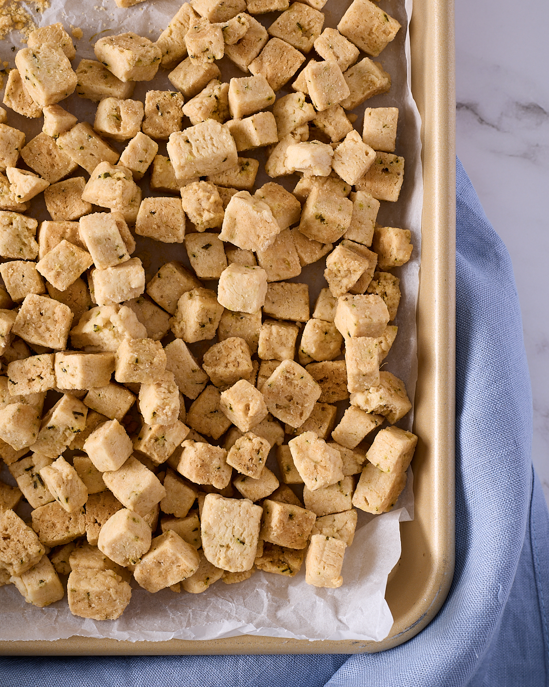 Tray of golden gluten free rosemary shortbread cubes cooling on baking paper, showing their light colour and crisp texture.