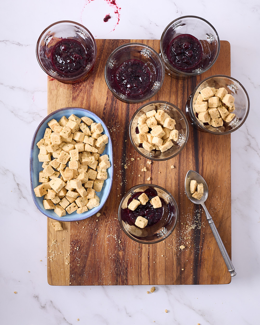 Overhead image of assembling blueberry coconut mini trifles, showing glass cups being layered with blueberry rosemary jam, gluten free rosemary shortbread cubes, and fresh blueberries on a wooden board.