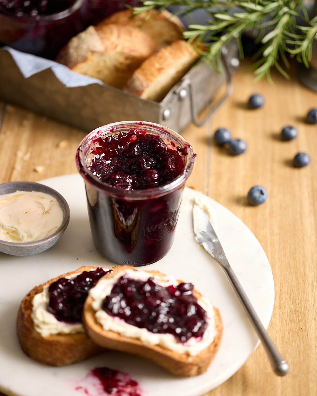 A jar of blueberry rosemary jam on a white plate with two slices of crusty bread spread with butter and jam, surrounded by fresh blueberries, rosemary sprigs, and a small bowl of butter.