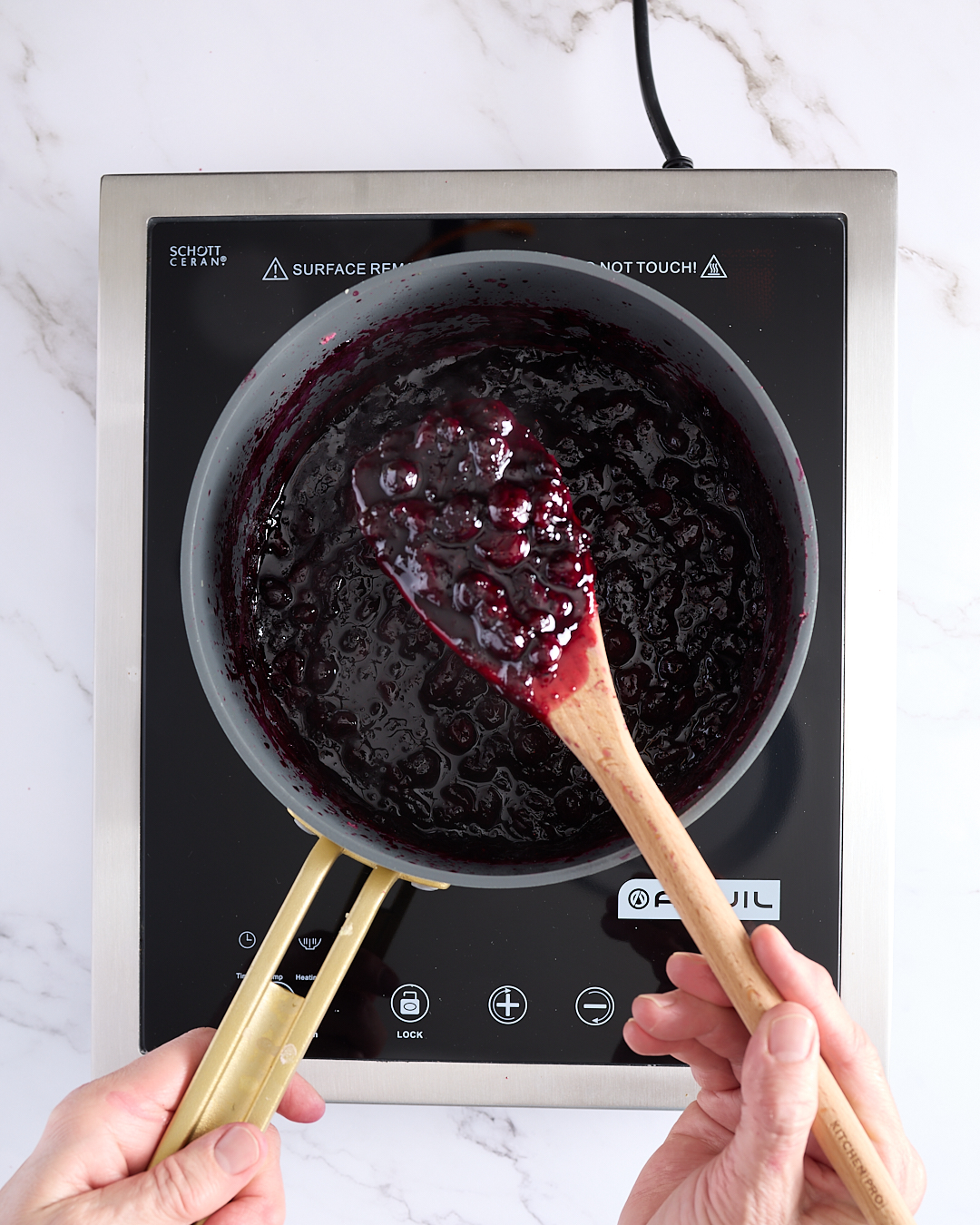 Overhead view of thick, glossy blueberry rosemary jam in a stainless-steel pot on an induction cooktop, with a wooden spoon lifting a scoop of the dark purple jam to show its texture and sheen.