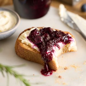 A slice of toasted sourdough spread with cream cheese and topped with blueberry rosemary jam, sitting on a marble plate beside a butter knife and jar of jam, with crumbs and blueberries scattered around.