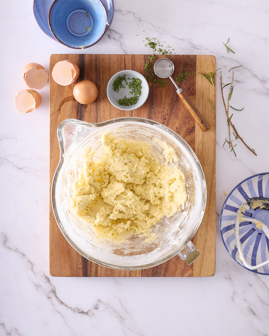 Mixing bowl of rosemary shortbread dough made with gluten free flour and coconut oil, shown on a wooden board with rosemary sprigs, eggshells, and small bowls of chopped rosemary and flour nearby.