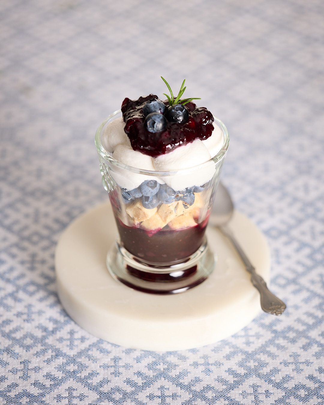 Single serve of blueberry coconut mini trifle in a glass, layered with blueberry rosemary jam, gluten free rosemary shortbread cubes, fresh blueberries, and whipped coconut yoghurt, topped with extra jam, blueberries, and a small sprig of rosemary, served on a white marble coaster with a spoon.