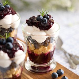 Close-up of individual blueberry coconut mini trifles layered in glass cups with blueberry rosemary jam, gluten free rosemary shortbread, fresh blueberries, and coconut whipped cream, topped with extra jam, blueberries, and a sprig of rosemary.