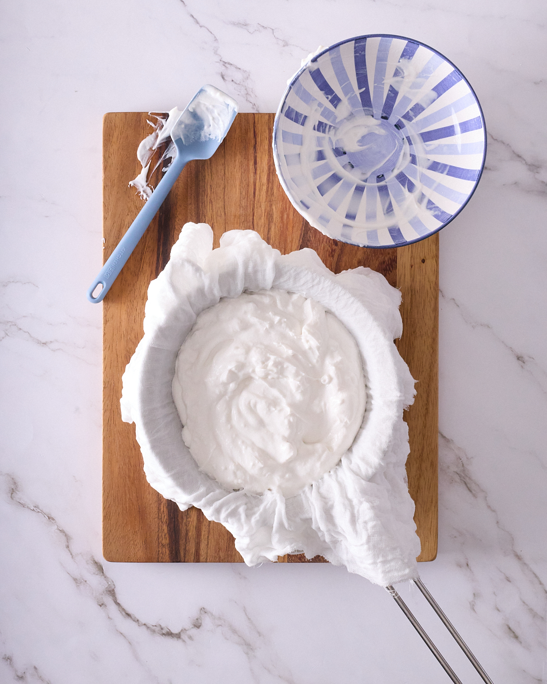 Straining coconut yoghurt through cheesecloth set over a bowl, showing the setup with spatula, empty mixing bowl, and yoghurt resting in the lined sieve ready to chill overnight.