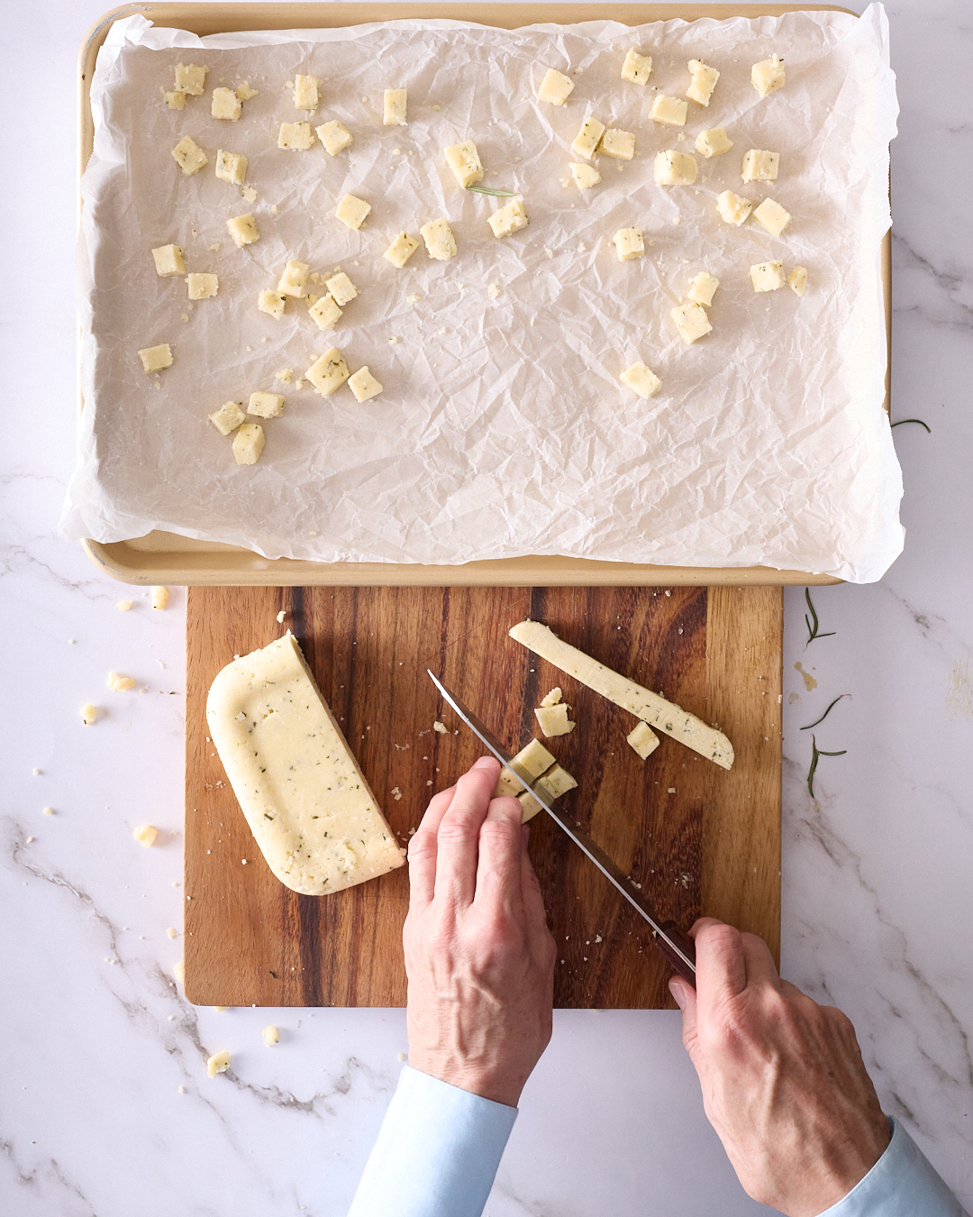 Sandra cutting chilled rosemary shortbread dough into small cubes on a wooden board, with cubed dough scattered on a parchment-lined baking tray ready for baking.