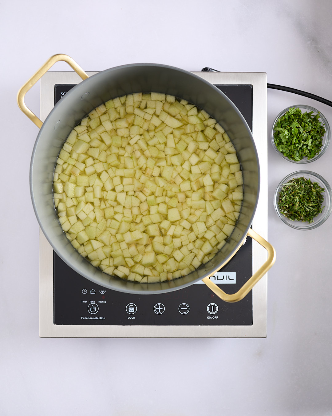 Diced Granny Smith apples in a large pot of liquid on an induction cooktop, with bowls of chopped mint and chopped rosemary beside the pot.