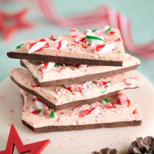 Stack of Candy Cane Peppermint Chocolate Bark on a white plate, layered dark and white chocolate studded with crushed red, white and green candy canes, with Christmas decorations and pinecones in the background.