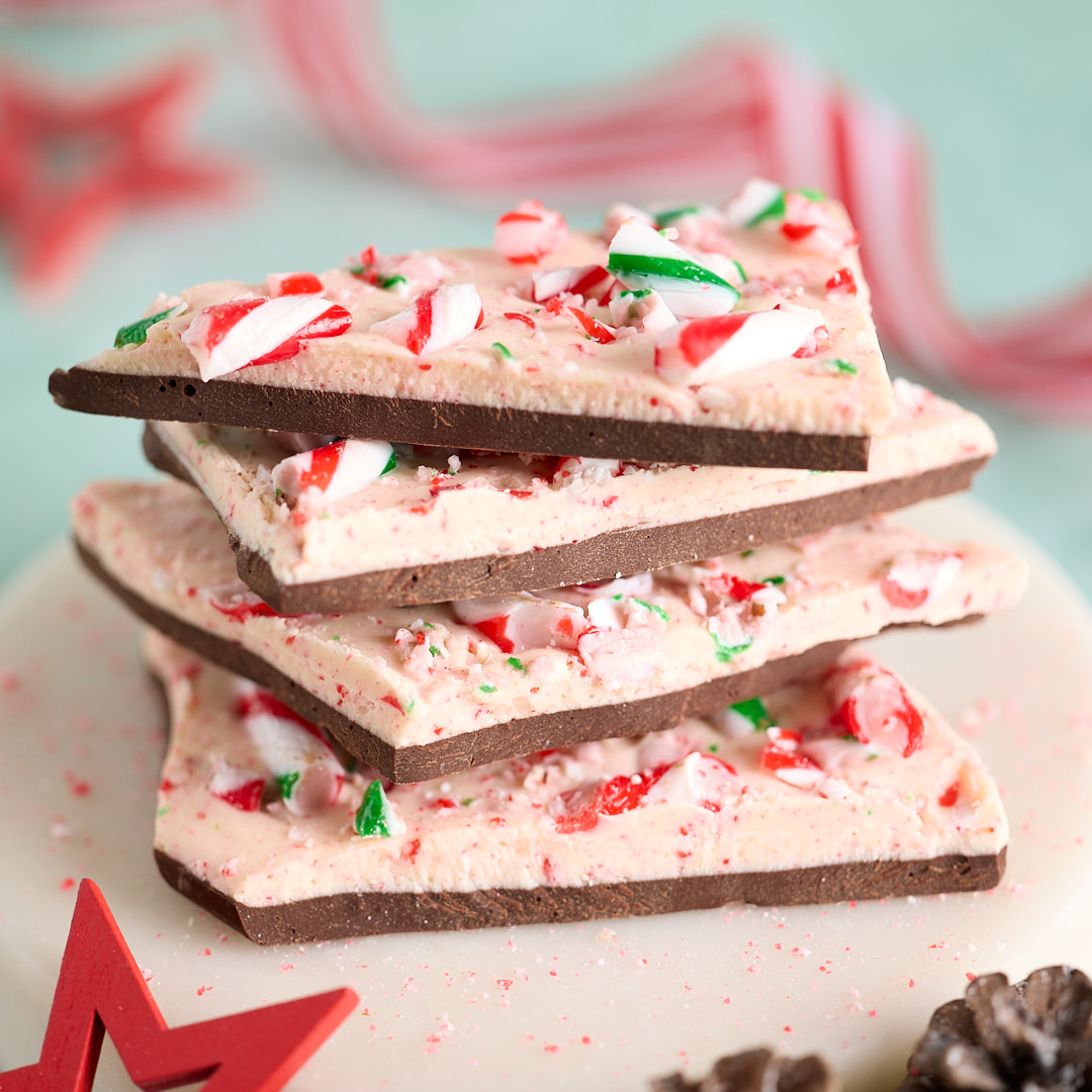 Stack of Candy Cane Peppermint Chocolate Bark on a white plate, layered dark and white chocolate studded with crushed red, white and green candy canes, with Christmas decorations and pinecones in the background.