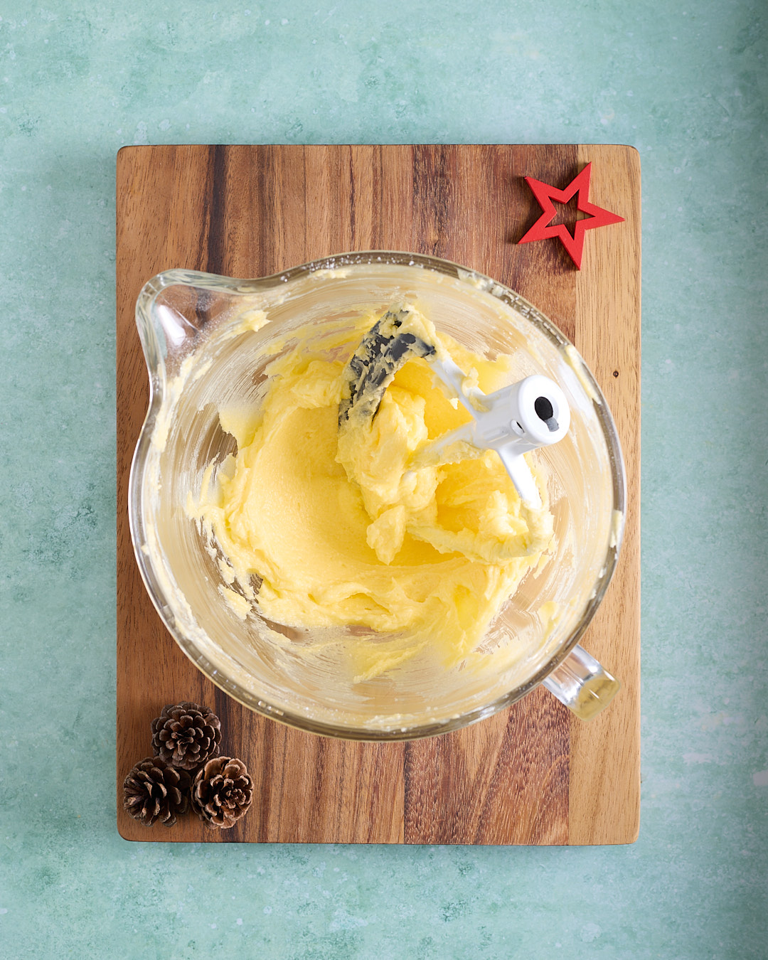 Overhead shot of a stand mixer bowl with butter and sugars beaten to a glossy pale yellow, with the paddle attachment resting in the bowl, styled on a wooden board with pinecones and a red star on a turquoise background.