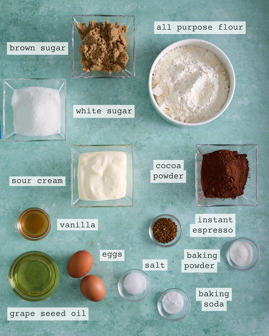Overhead flat lay of the labelled ingredients for devil’s food mini cakes on a teal background: all-purpose flour, Dutch processed cacao powder, brown sugar, white sugar, sour cream, vanilla, grapeseed oil, eggs, instant espresso, salt, baking powder, and baking soda in small bowls.