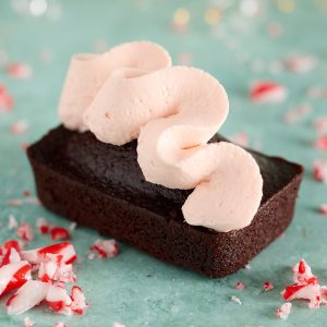Close-up of a devil’s food mini cake topped with a swirl of whipped peppermint bark ganache, with crushed candy cane pieces scattered on a teal background.