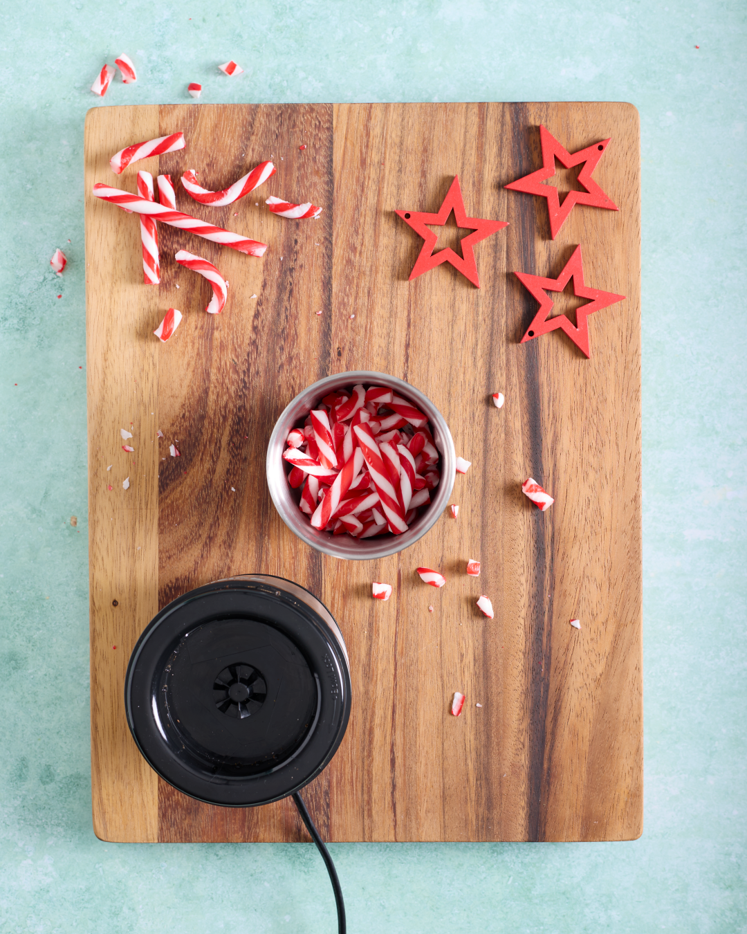 Overhead image of whole and broken red-and-white candy canes on a wooden board, with pieces collected in a small metal cup next to a black electric spice grinder and red star decorations.