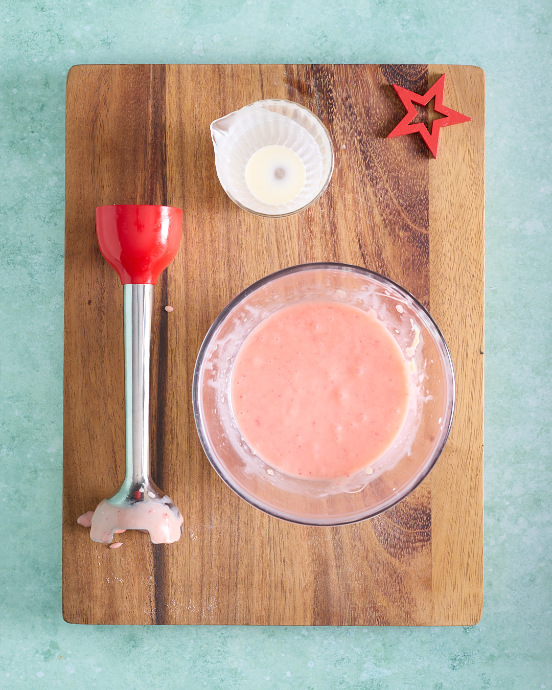 Overhead image of pale pink candy cane white chocolate ganache mixture in a glass bowl on a wooden board, with a stick blender and a small jug nearby.