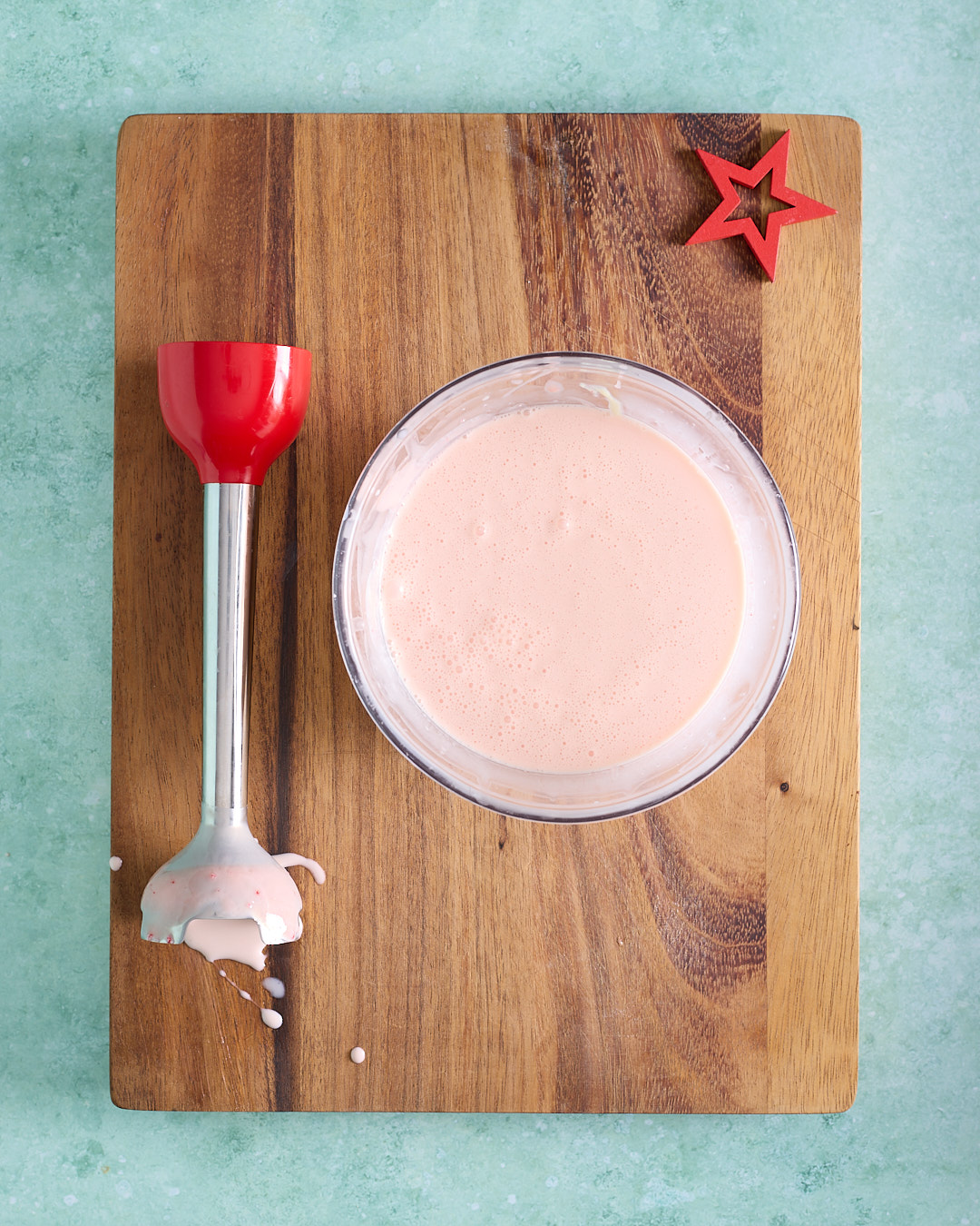 Overhead image of smooth, pale pink candy cane white chocolate ganache mixture in a glass bowl on a wooden board, with a stick blender beside it.